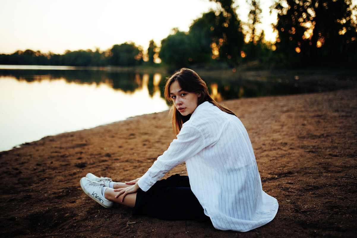 Девушка в белой рубашке и черных брюках сидит на песчаном берегу реки во время заката в Уфе - Lenar Abdrakhmanov
