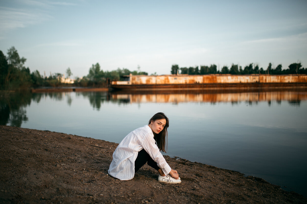 Девушка в белой рубашке и черных брюках сидит на песчаном берегу у реки с заброшенным кораблем - Lenar Abdrakhmanov