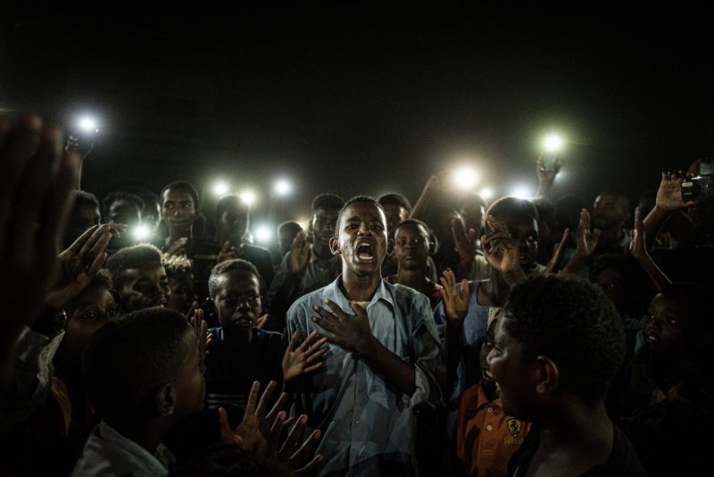 «Фото года», 2020. Участник протестов против режима Омара аль-Башира читает стихи в свете телефонов при отключенной электроэнергии в Хартуме, Судан. Автор Ясуёси Тиба.